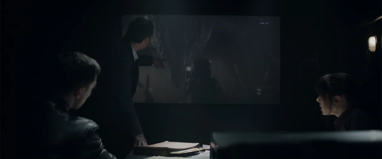 Godzilla (2014) movie still: medium — A dark, dimly lit conference room contains a large projection screen on the wall. A man stands before the…