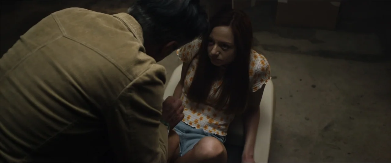 Captive (2020) movie still: medium — A dimly lit concrete basement contains a man and a young woman. The man stands over the woman who is seated on…