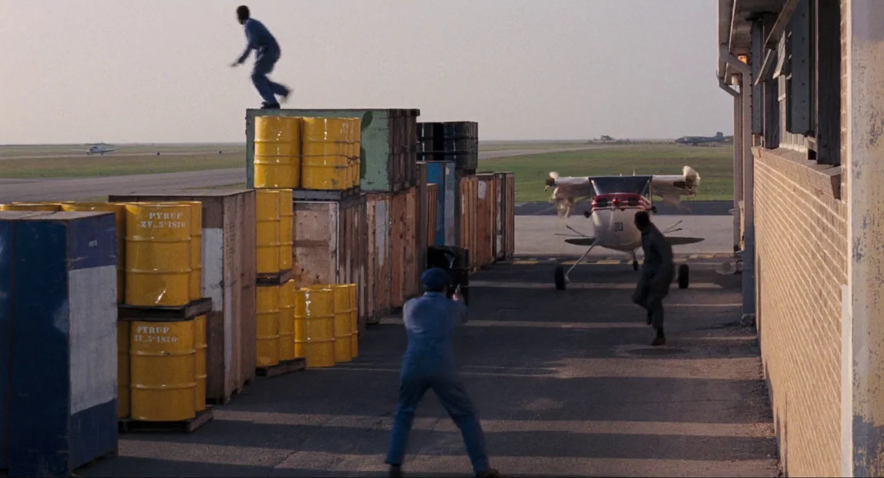 Live and Let Die (1973) movie still: wide — An open airfield at midday with stacked yellow barrels and wooden crates near hangars. Three men in work…