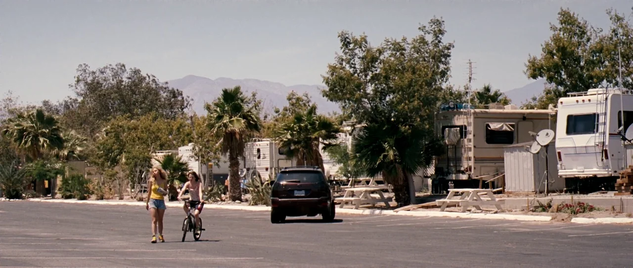 Little Birds (2011) movie still: wide — A sunny RV park lot features several large travel trailers and palm trees. A woman walking and a girl on a…