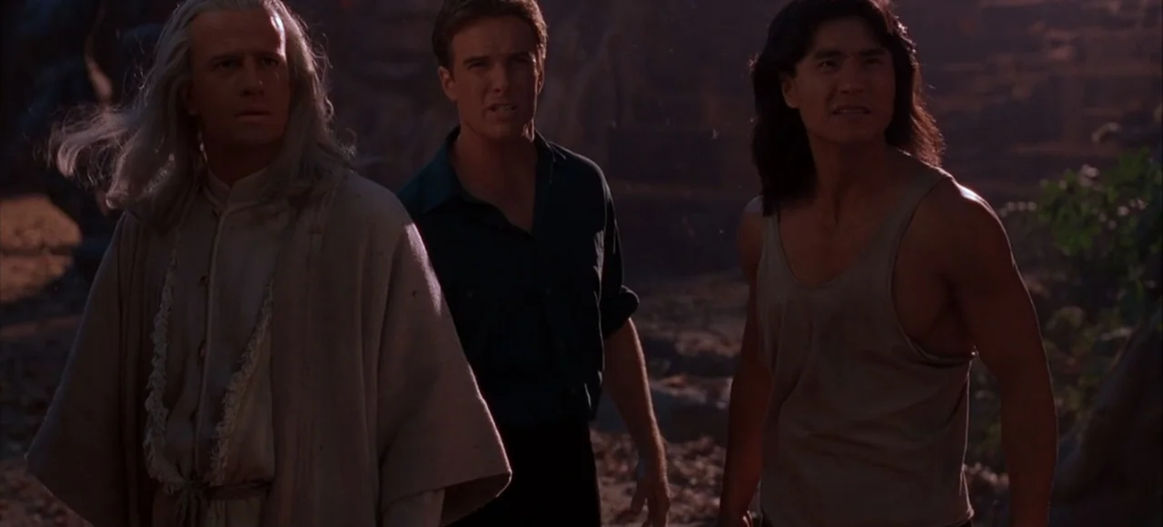 Mortal Kombat (2021) movie still: medium — Three men stand on a rugged outdoor surface during the late afternoon. They are positioned side-by-side…