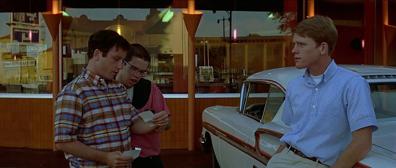 American Graffiti (1973) movie still: medium — A 1960s style restaurant exterior with large glass windows and orange structural pillars. Three young…