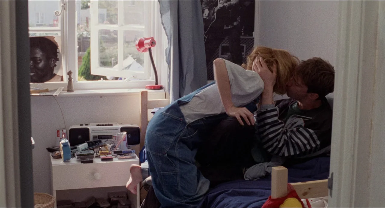 Life Is Sweet (1990) movie still: medium — A cluttered 1990s bedroom with a white side table and a window looking outside. A young man and woman in…