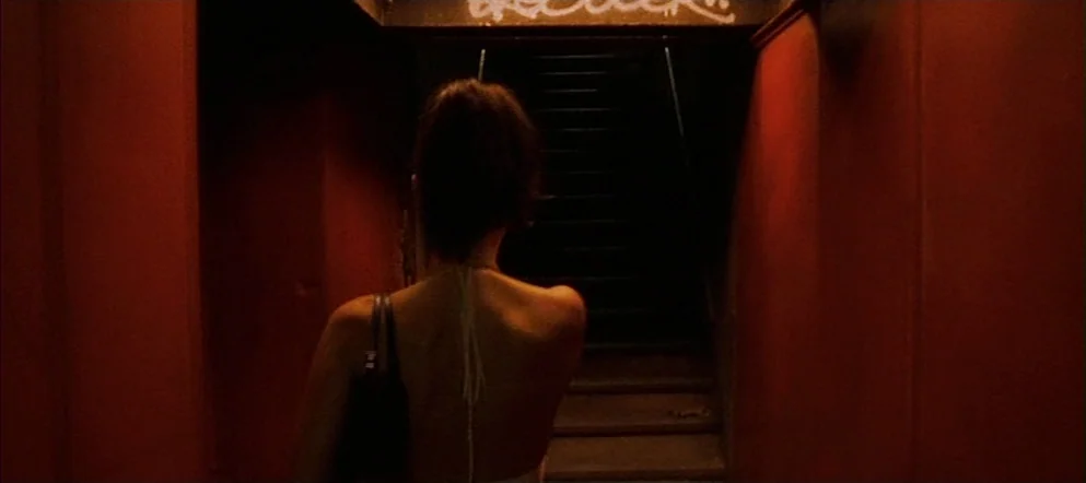 Irréversible (2002) movie still: medium — A narrow stairwell with deep red walls and dark, shadowed steps leading upward. A woman with dark hair in…