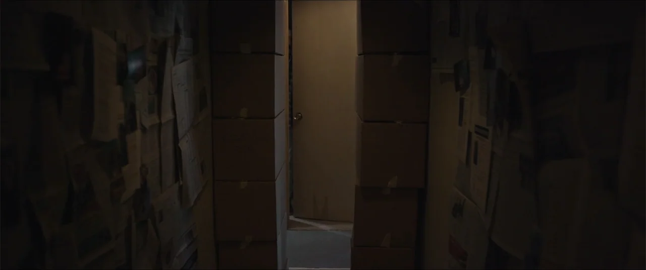 Captive (2020) movie still: medium — A narrow interior corridor is flanked by high stacks of brown cardboard moving boxes. The walls are covered…
