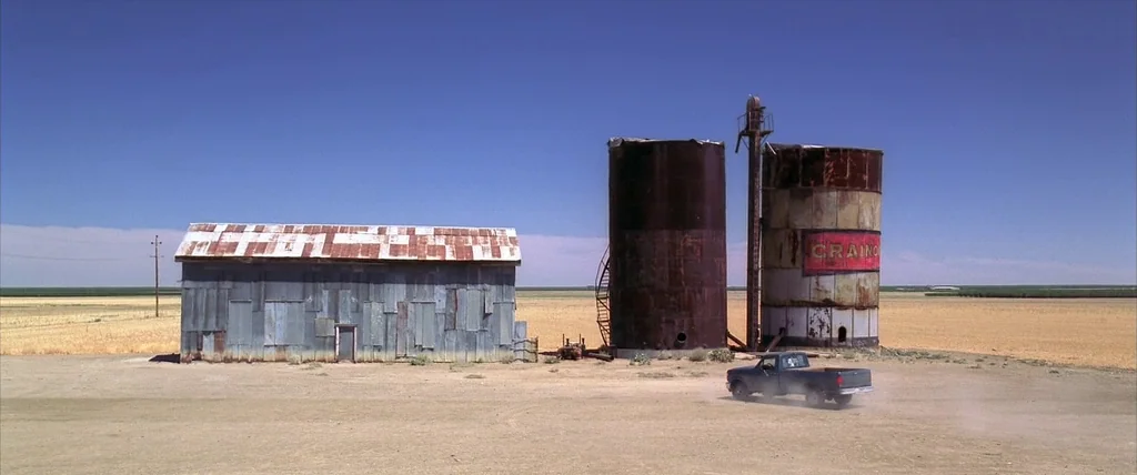 The Cell (2000) movie still: wide — A desolate farm lot features a metallic shed and two weathered grain silos. A small dark pickup truck kicks up…