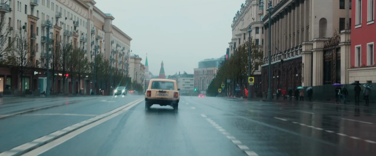 Anna (2019) movie still: wide — A wide, multi-lane urban street in Moscow is wet from recent rainfall. A beige Lada hatchback drives away from the…