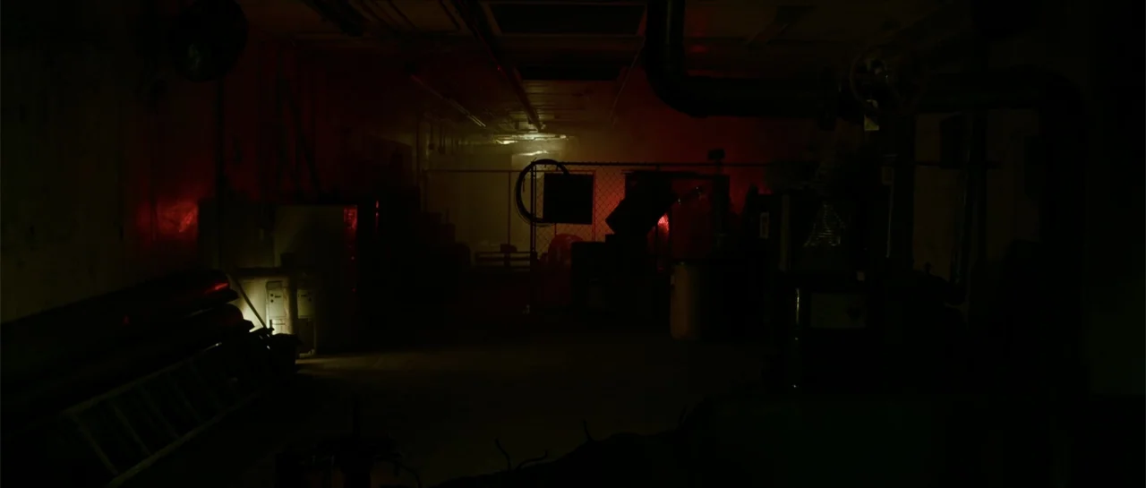 The Vault (2017) movie still: establishing — A cluttered, dark interior space features exposed overhead pipes and a chain-link fence boundary. No…