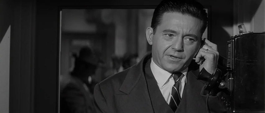 Compulsion (1959) movie still: medium — A man stands inside a dimly lit booth using a wall-mounted telephone handset. The background shows a blurry…