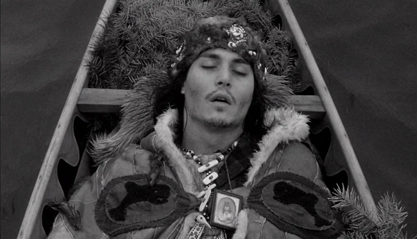 Dead Man (1995) movie still: medium — A wooden canoe filled with pine branches sits under diffuse daylight. A young man lies with eyes closed in the…
