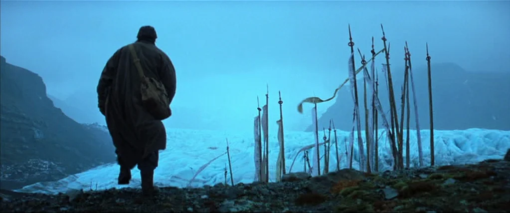 Batman Begins (2005) movie still: wide — A vast glacier sits between dark mountain slopes under a thick, overcast sky. A person wearing a long coat…