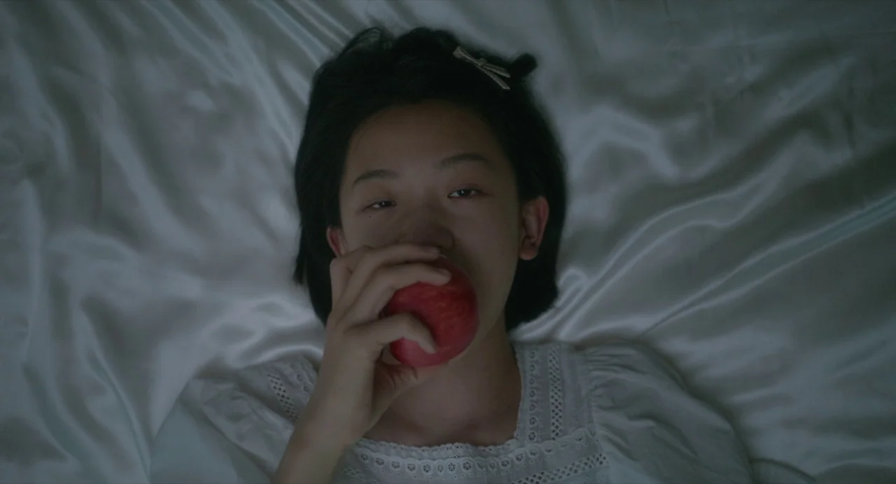 White River (2023) movie still: closeup — A person lies flat on white satin sheets beneath an overhead light source. The subject rests on the fabric…