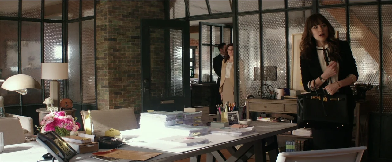 Fifty Shades Freed (2018) movie still: medium — A sunlit office space features brick walls, glass partitions, and a large wooden desk. A woman with…