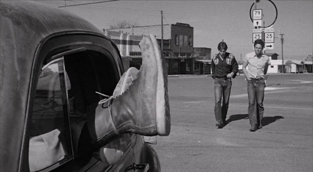 The Last Picture Show (1971) movie still: medium — An empty, small-town intersection with aged buildings and a prominent road sign. Two men walk…