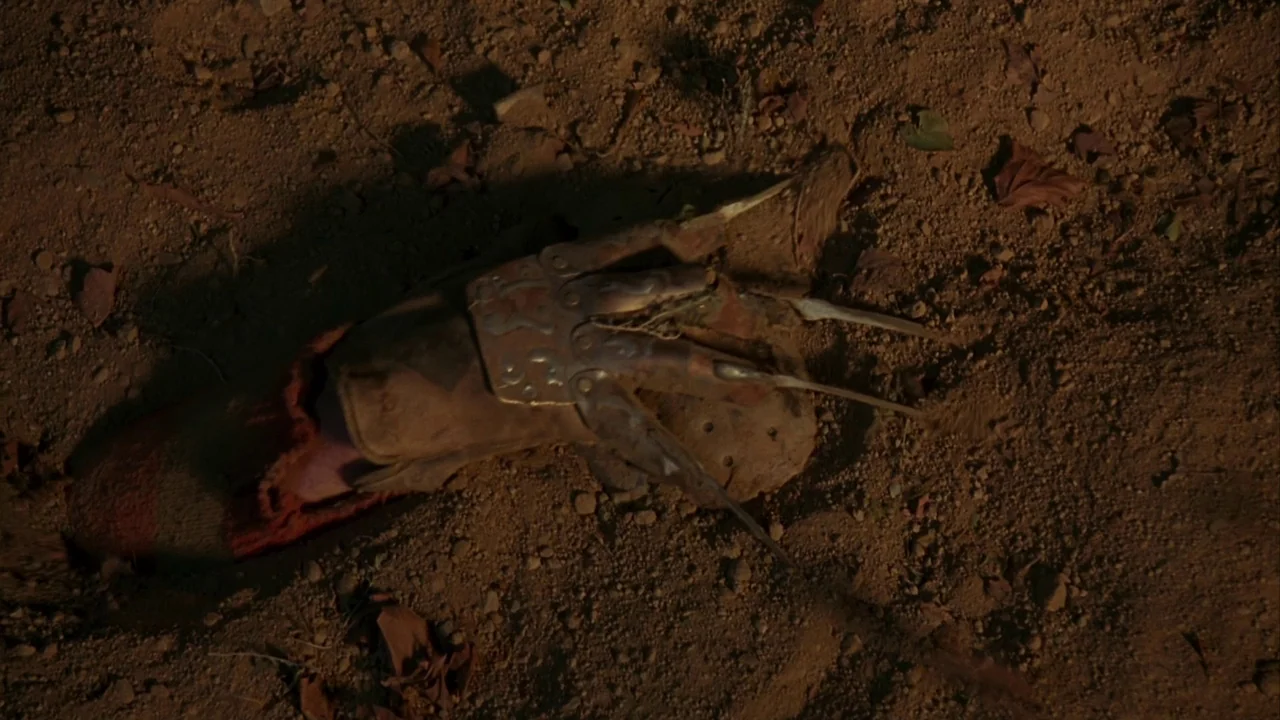 Jason Goes To Hell (1993) movie still: An ominous close-up frames a notorious, clawed metal glove resting on dark, reddish-brown earth, scattered…