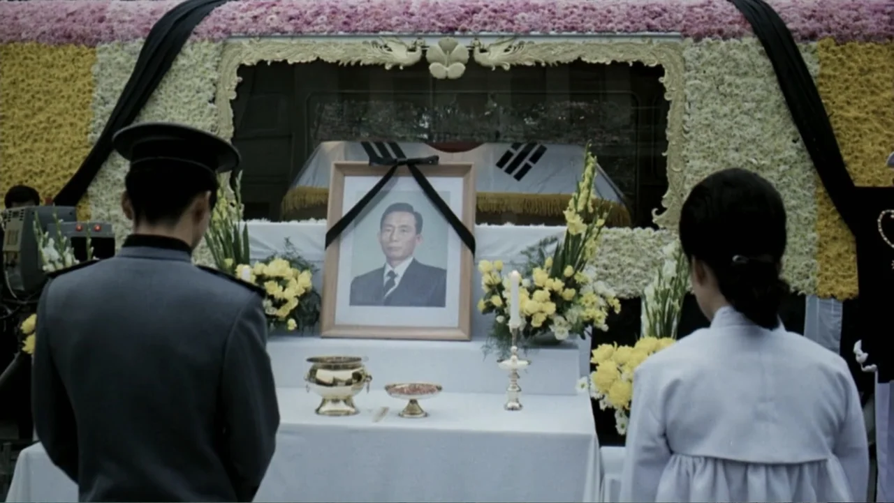 Lee Soo Man: King of K-Pop (2025) movie still: medium — A formal funerary altar features a portrait photograph surrounded by yellow and white…