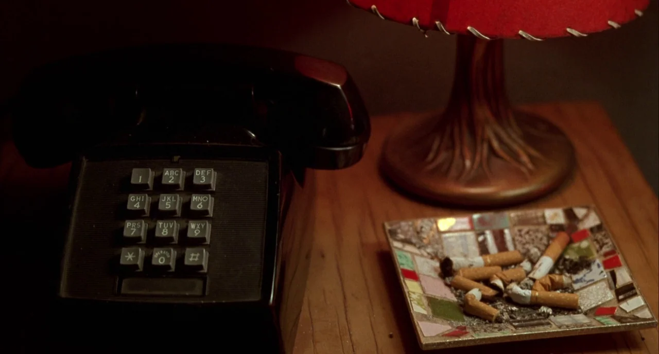 Mulholland Drive (2001) movie still: closeup — A wooden nightstand surface holds a black telephone, a lamp, and a mosaic ashtray. Several cigarette…