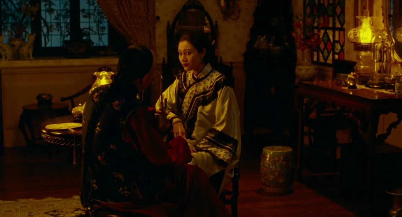 Flowers of Shanghai (1998) movie still: medium — A dimly lit room with vintage decor includes a table lamp and patterned textiles. A woman in a…