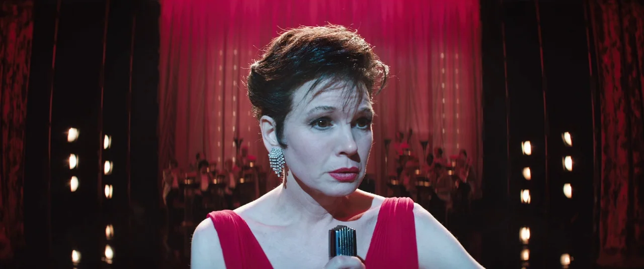 Judy (2019) movie still: closeup — A dark concert stage featuring vertical rows of illuminated practical lights and red curtains. A woman in a red…
