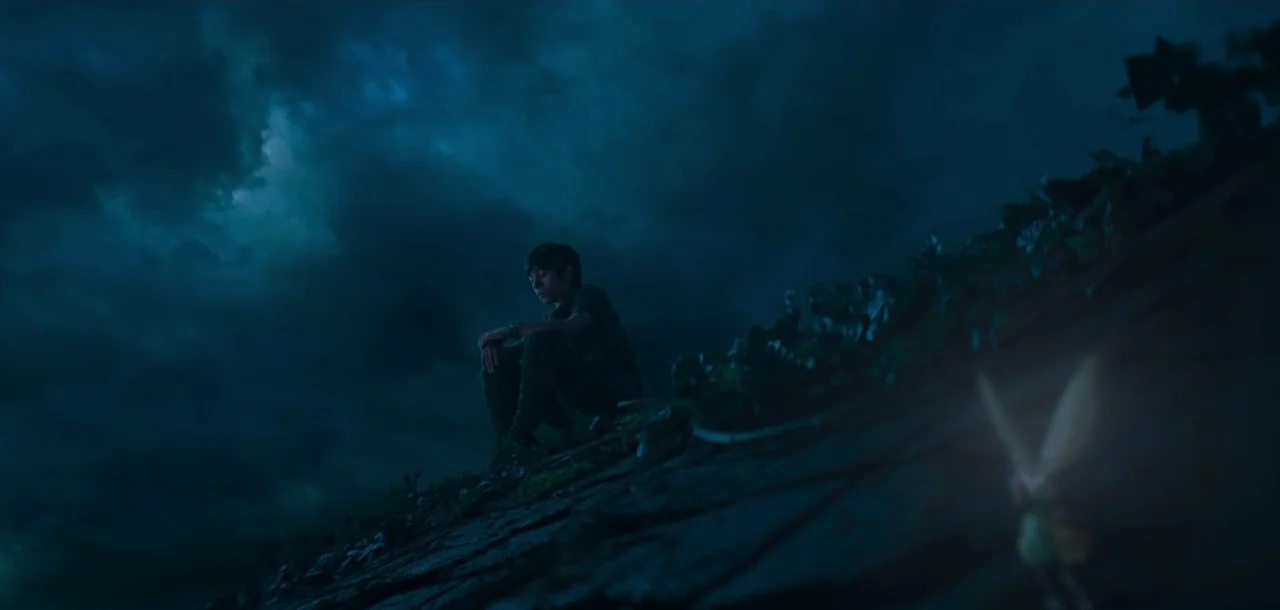 Peter Pan & Wendy (2023) movie still: wide — A dark night sky looms over a steeply pitched, dark tiled roof. A boy sits alone on the tiles as a…