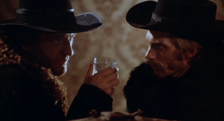 Roy Colt and Winchester Jack (1970) movie still: medium — A dimly lit saloon interior features two men seated opposite each other in shadows. They…