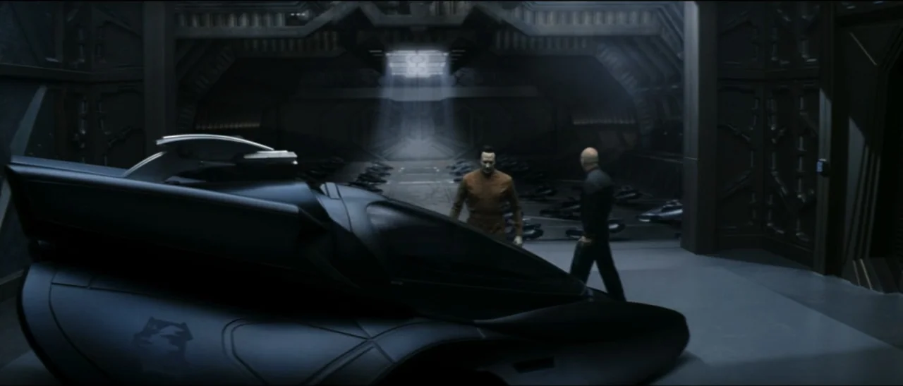 Star Trek: Nemesis (2002) movie still: wide — A spacious dark hangar features a sleek black spacecraft parked in the foreground. Two men walk…