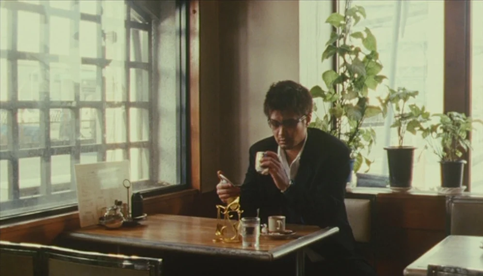 Gozu (2003) movie still: medium — A quiet corner of a restaurant featuring a wooden table and vinyl-covered bench seats. A man sits by the window,…
