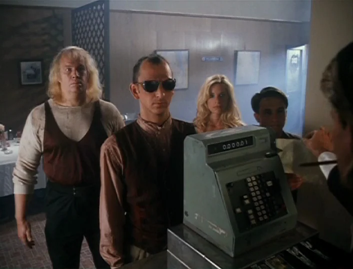 Head of the Family (1996) movie still: medium — A dated motel interior with wood-paneled walls and a beige cash register. Four people stand before…