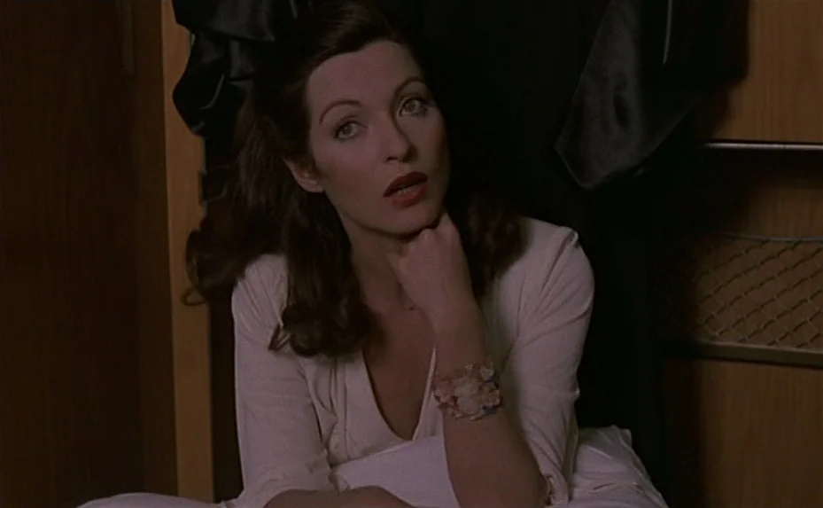 Love on the Run (1979) movie still: medium — A dark bedroom with wood-paneled walls and a draped black garment in the background. A woman in a light…