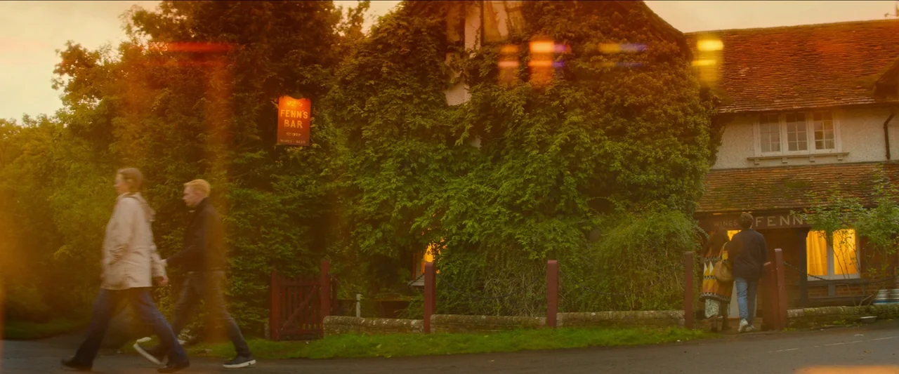 Unwelcome (2022) movie still: wide — A historic timber-framed pub covered in dense ivy stands behind a low stone wall. Two people walk along the…