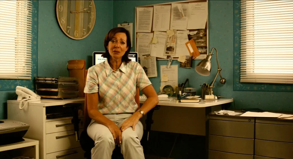 Life During Wartime (2009) movie still: medium — A small room has blue wallpaper covered with documents pinned to a bulletin board. A woman sits in…