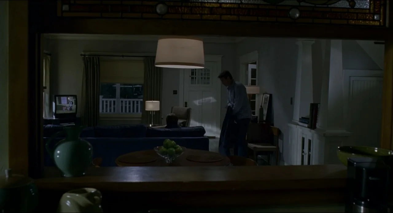 The Ring Two (2005) movie still: medium — A dimly lit living room viewed through a kitchen pass-through window at night. One man stands by the front…