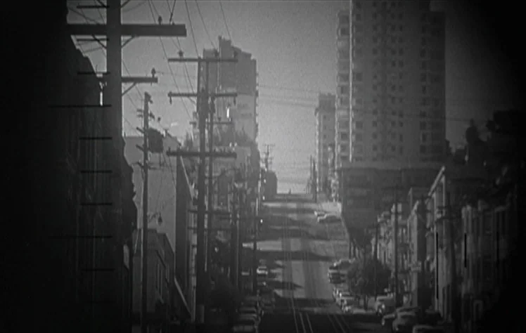 On The Beach (1959) movie still: establishing — A steep residential street in a city is lined with utility poles and buildings. Parked cars are…