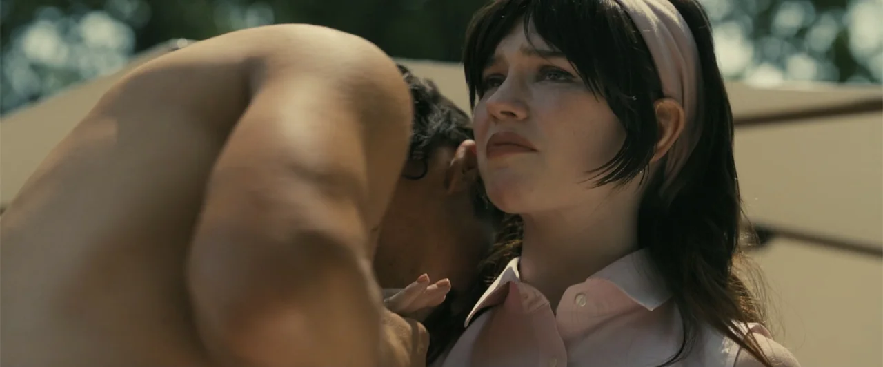 Companion (2025) movie still: medium — A campsite exterior during daylight with a man and woman in close proximity. The man stands shirtless beside…