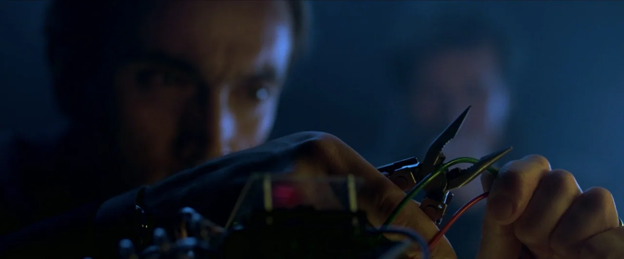 V for Vendetta (2006) movie still: closeup — A dark, dimly lit environment where an individual performs precise work on machinery. A man uses pliers…