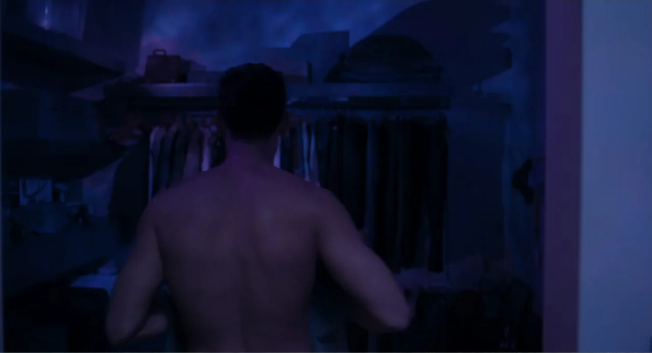 Straight (2024) movie still: medium — A dimly lit walk-in closet is filled with racks of dark hanging garments. A bare-chested man stands with his…