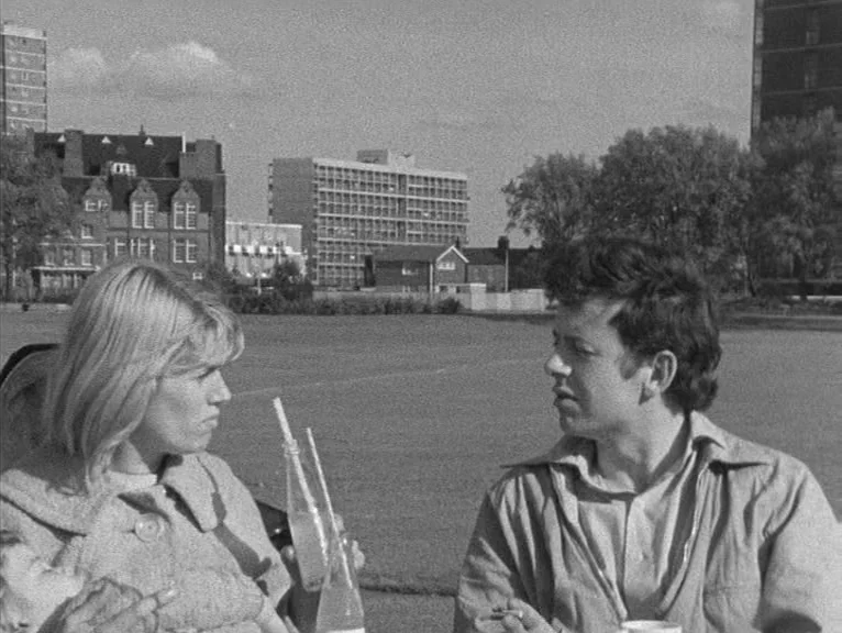 Cathy Come Home (1969) movie still: medium — An open park area with mid-century modern apartment buildings in the background. A young couple sits…