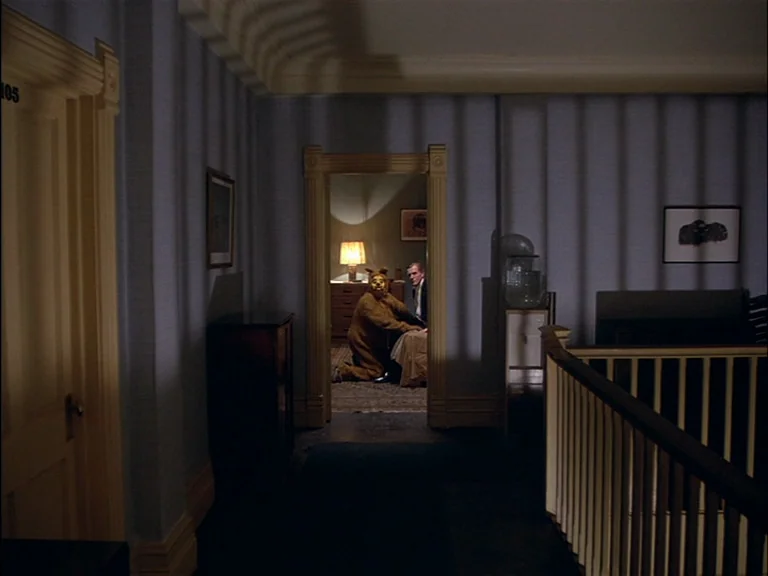 The Shining (1980) movie still: wide — A dimly lit hotel hallway features dark blue walls with sharp vertical shadow patterns. A person in a bear…