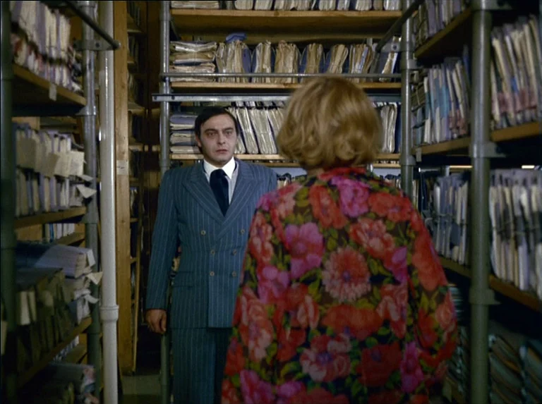 Satan’s Brew (1976) movie still: medium — An narrow aisle filled with metal shelves holding rows of paper files and documents. A man in a pinstriped…