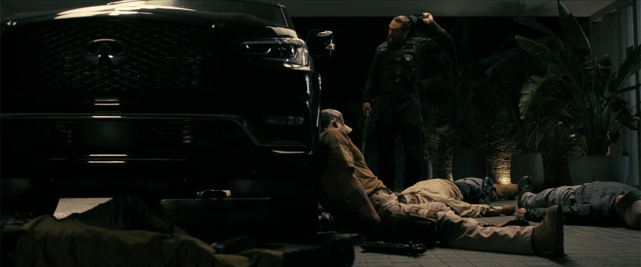 The Wrecking Crew (2026) movie still: wide — A modern dimly lit garage features a large black vehicle parked on concrete. One standing man adjusts…