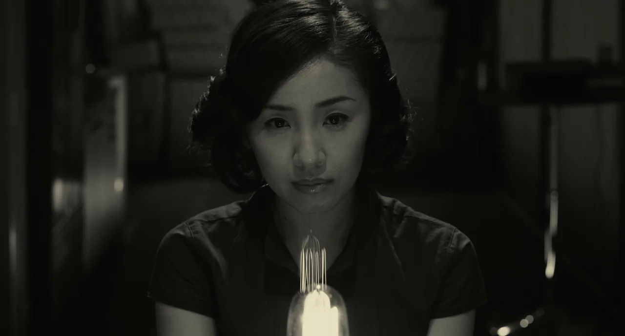 The Whispering Star (2015) movie still: closeup — A woman stands in a dimly lit, dark space with minimal background detail visible. She holds a…