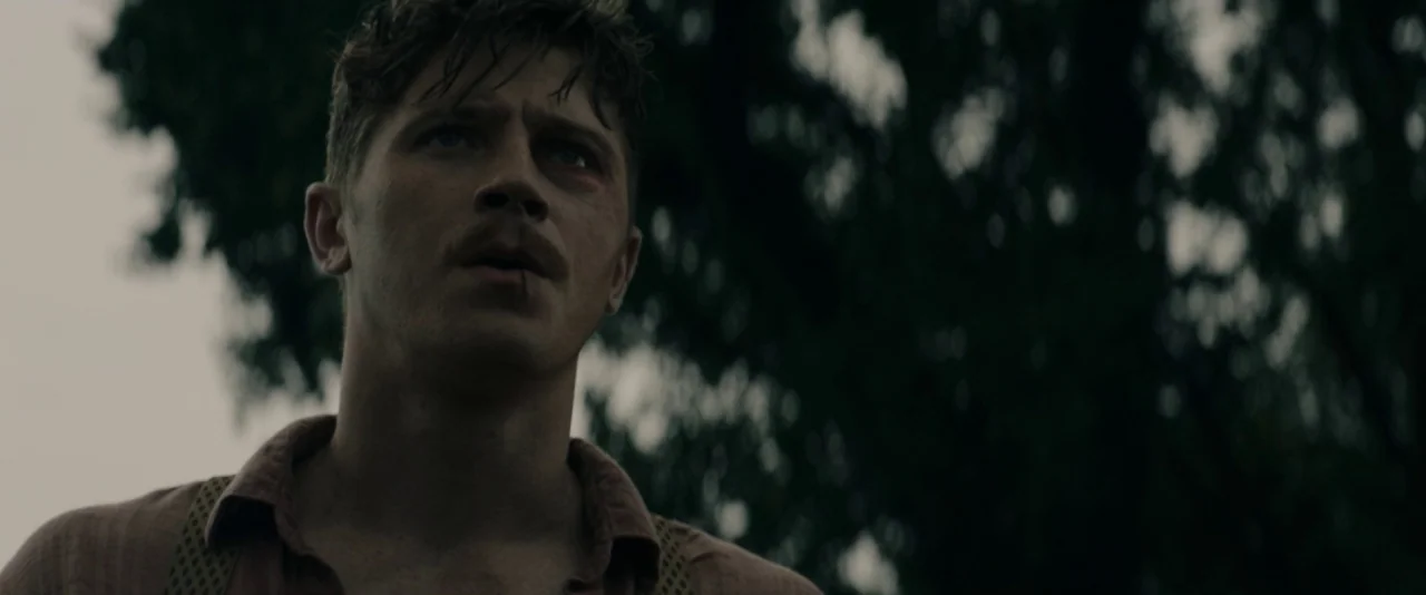 Mudbound (2017) movie still: medium — An outdoor area with blurred dark tree silhouettes against a gray overcast sky. A man looks upward with a…