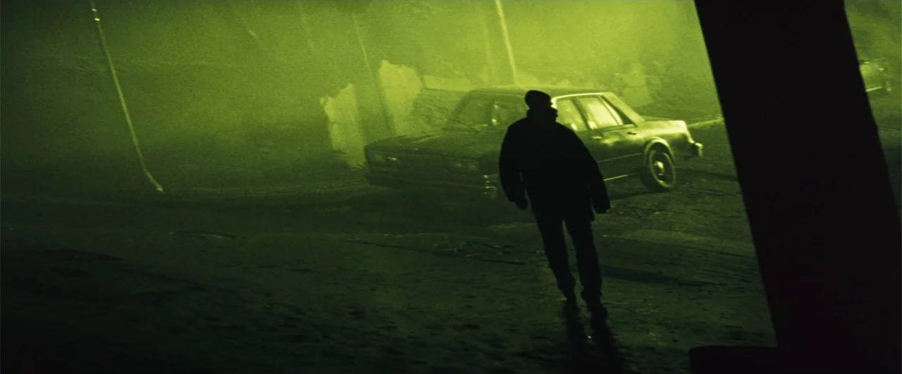 Fallen (2004) movie still: wide — A dark urban environment is filled with thick green fog and low light. A man walks across the foreground as an…