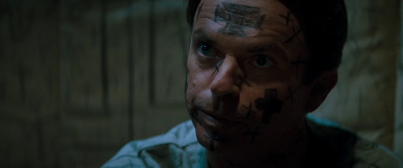 In The Mouth of Madness (1994) movie still: closeup — A dimly lit room with textured, dark walls surrounds the central subject. A man with distinct…