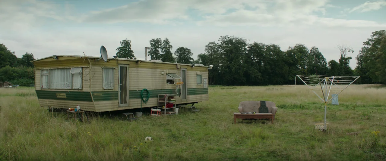 Mindhorn (2016) movie still: wide — A worn, light-colored mobile home sits in an overgrown meadow under a cloudy sky. Beside the home, an outdoor…