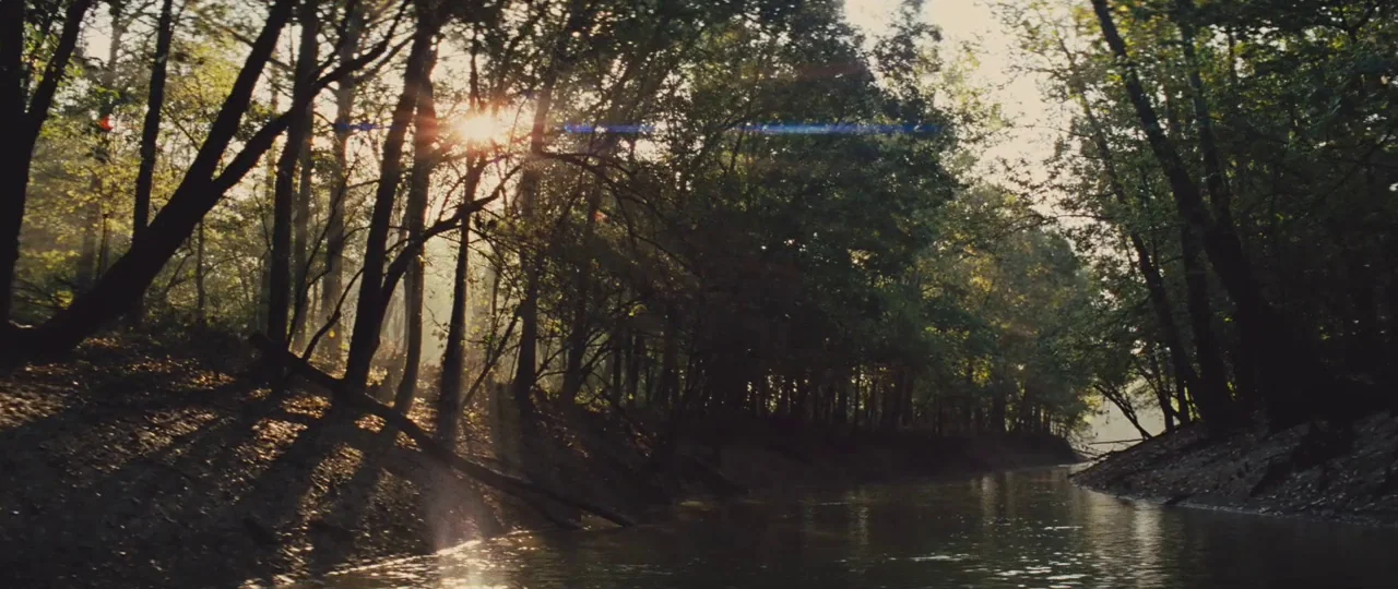 Mud (2012) movie still: establishing — A slow-moving muddy river winds through a dense temperate deciduous forest at midday. Sunlight cuts through…