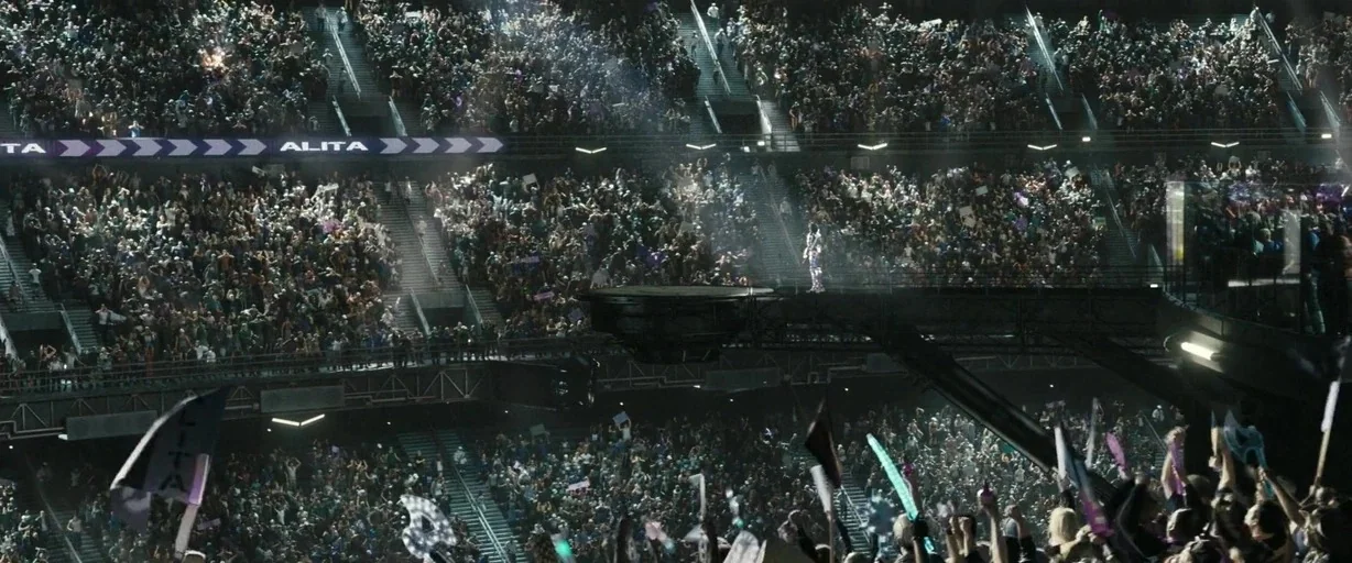 Alita: Battle Angel movie still: wide — A vast futuristic sports stadium filled with a dense crowd of cheering spectators. A solitary figure stands…