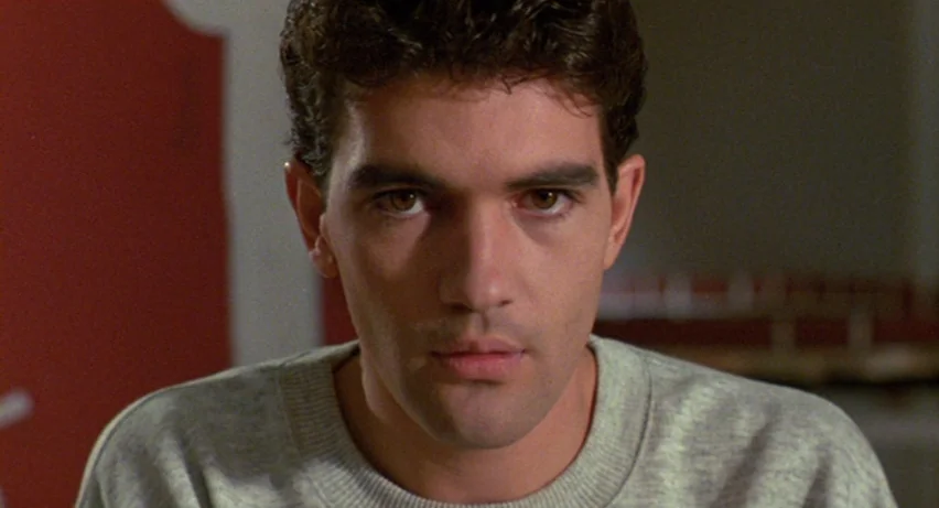 Matador (1986) movie still: closeup — An interior space features a solid red wall behind a young man. The man stares directly into the camera lens…