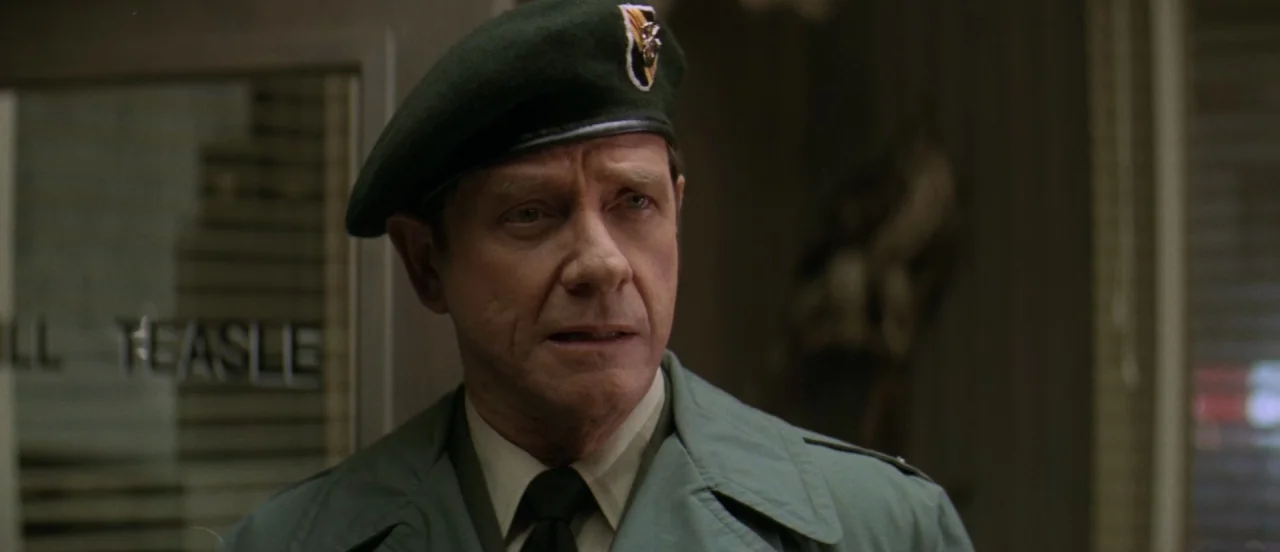 First Blood (1982) movie still: closeup — A man in a green beret and uniform stands inside an office setting. Text on a glass door behind him reads…