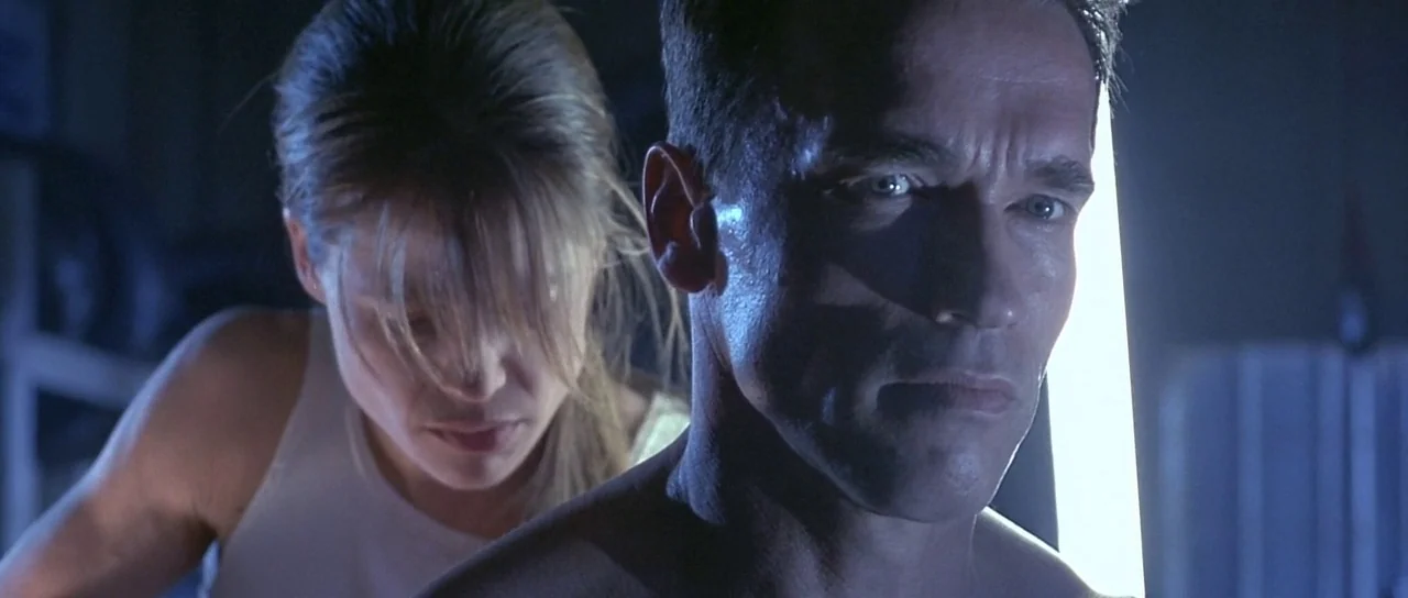 Terminator 2: Judgement Day (1991) movie still: medium — A dimly lit industrial space with a bright light source behind a male figure. A woman…